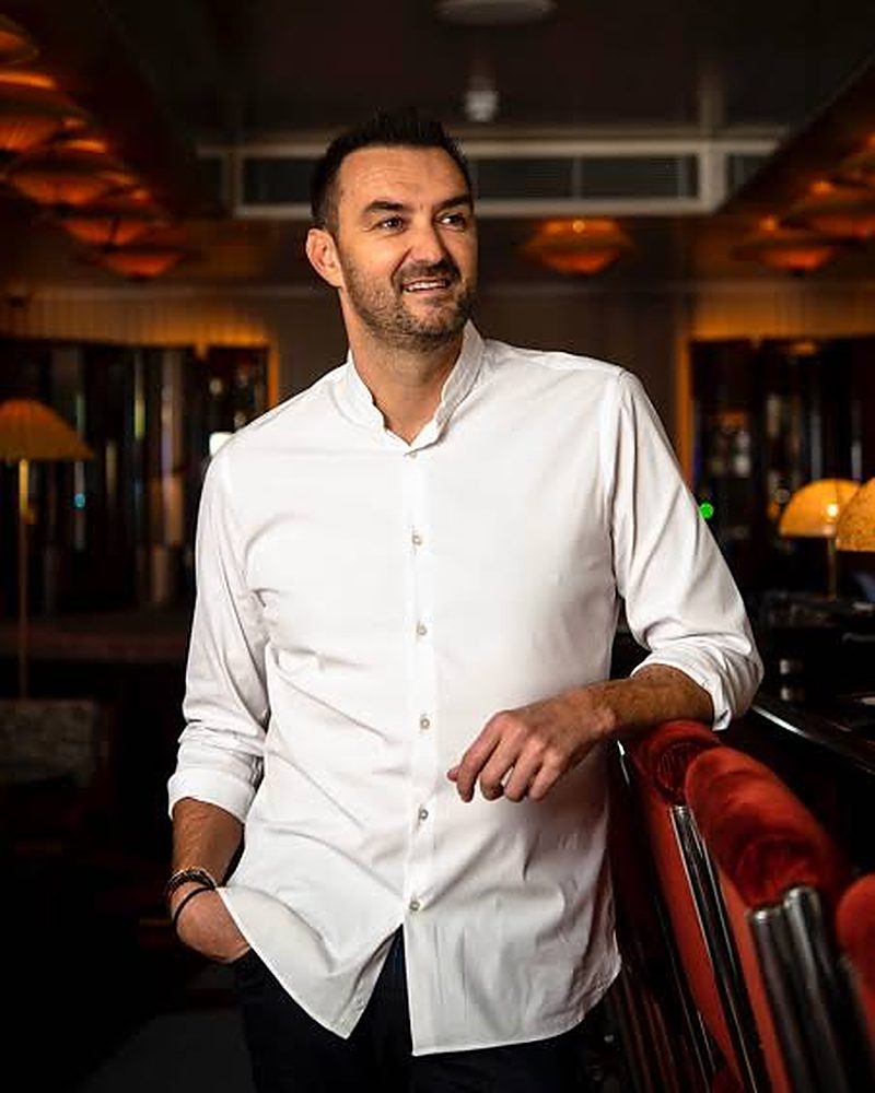 Cyril Lignac, chef, en chemise blanche, accoudé à la banquette rouge de la salle de son restaurant