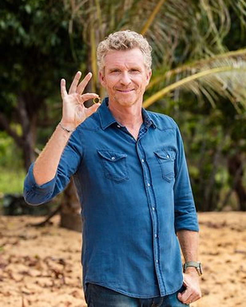 Denis Brogniart, animateur de Koh-Lanta, en chemise en jean, l’anneau d’or entre les doigts, sur la plage de l’aventure